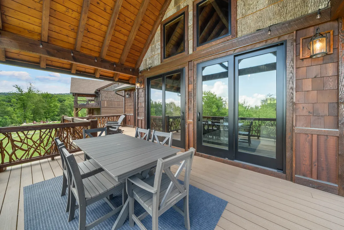 A spacious wooden outdoor dining table is set on a deck, framed by leafy greenery and large glass doors that lead back inside. The view extends to the surrounding landscape, and the mood is tranquil—ideal for sunrise coffee, al fresco meals, or simply soaking in the mountain setting. The image invites viewers to imagine themselves relaxing outdoors, making the most of the deck’s scenic positioning.