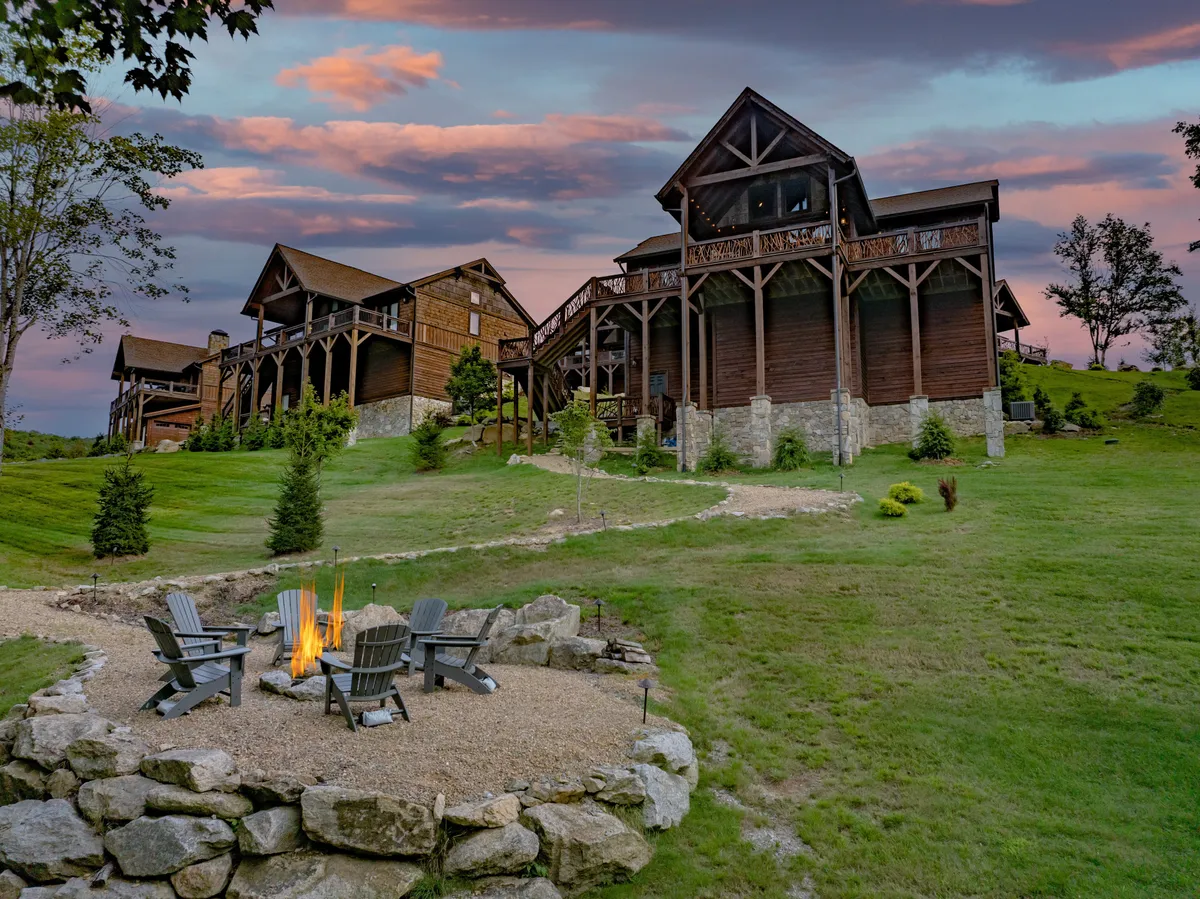 A wide view of the backyard reveals a fire pit surrounded by outdoor seating, a large wooden cabin, and a sweeping mountain landscape beyond. The setup is perfect for campfire gatherings, outdoor dining, laughter, and enjoying the natural surroundings together.
