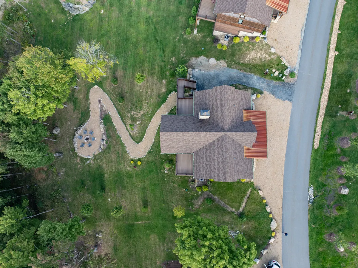 A serene exterior view of a winding pathway leading to a central fire pit area, with inviting seating softened by surrounding greenery. The roof of a nearby house peeks out in the background, and the mood is calm and welcoming, suggesting opportunities for memorable outdoor gatherings.