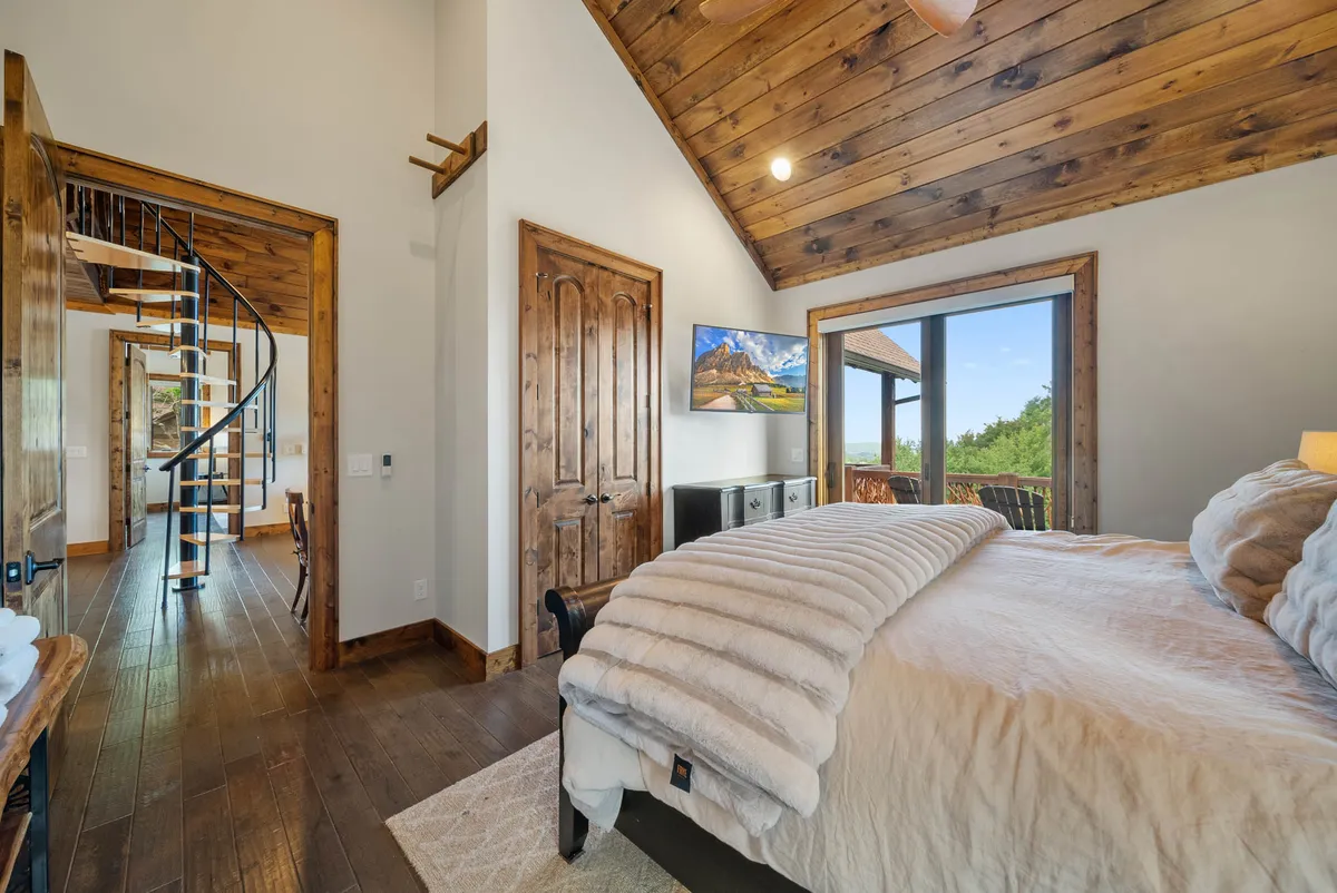 A spacious bedroom with plush bedding and a large window offering a scenic view of the outdoors. The spiral staircase and the hint of the deck outside suggest easy access to fresh air and outdoor spaces.