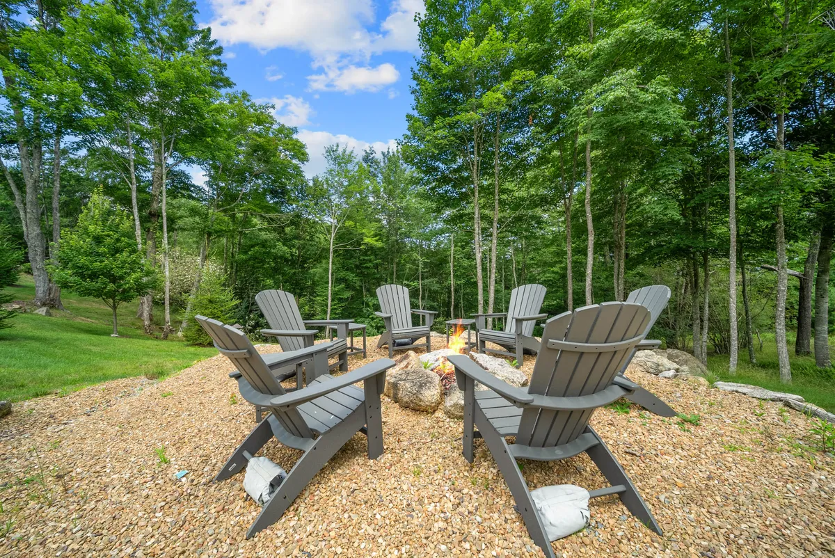 A cozy fire pit area is nestled among trees and surrounded by adirondack chairs. The gravel area enhances the rustic, natural vibe, and the scene is clearly designed for groups to gather, relax, and connect. The setting is communal and inviting, perfect for campfire storytelling, making s'mores, or stargazing under a mountain sky.