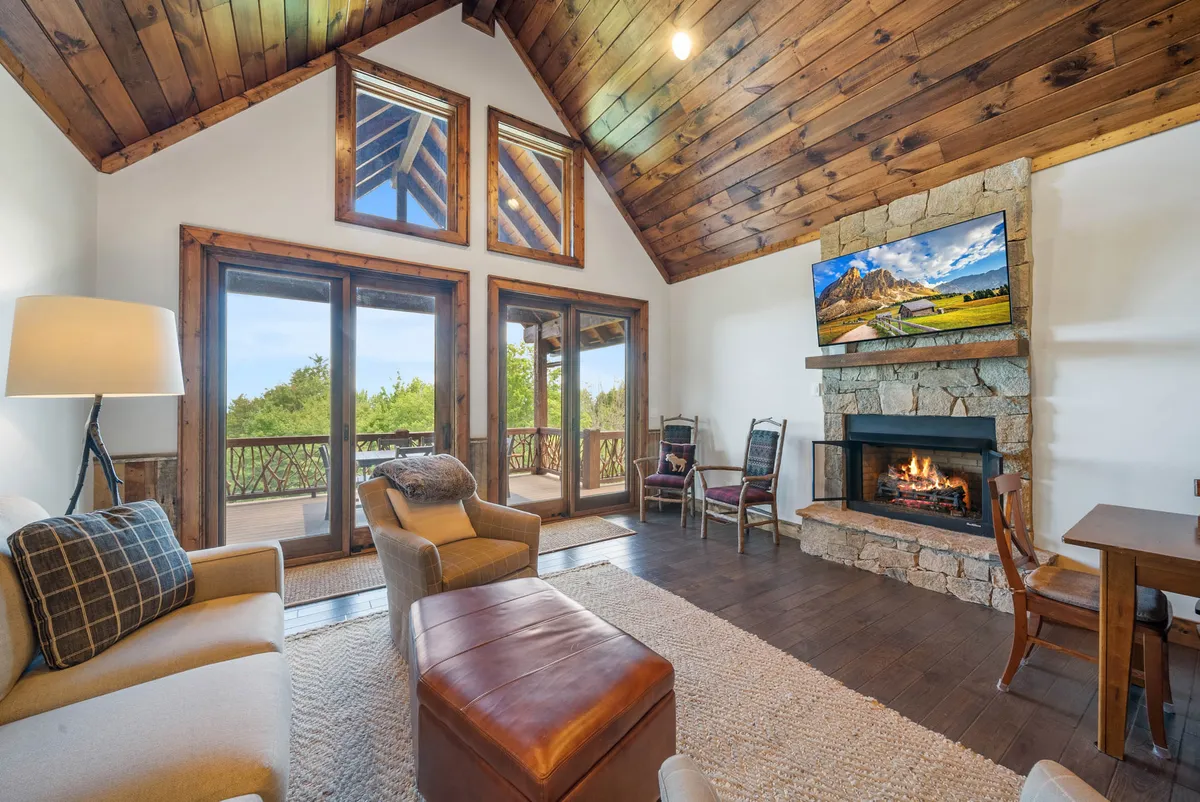 This image showcases the main living area with plush, comfortable seating arranged around a cozy gas fireplace. Large windows reveal sweeping mountain vistas, while exposed wooden beams add warmth and rustic character. A television is visible, perfect for movie nights, and the space exudes a welcoming, communal atmosphere.