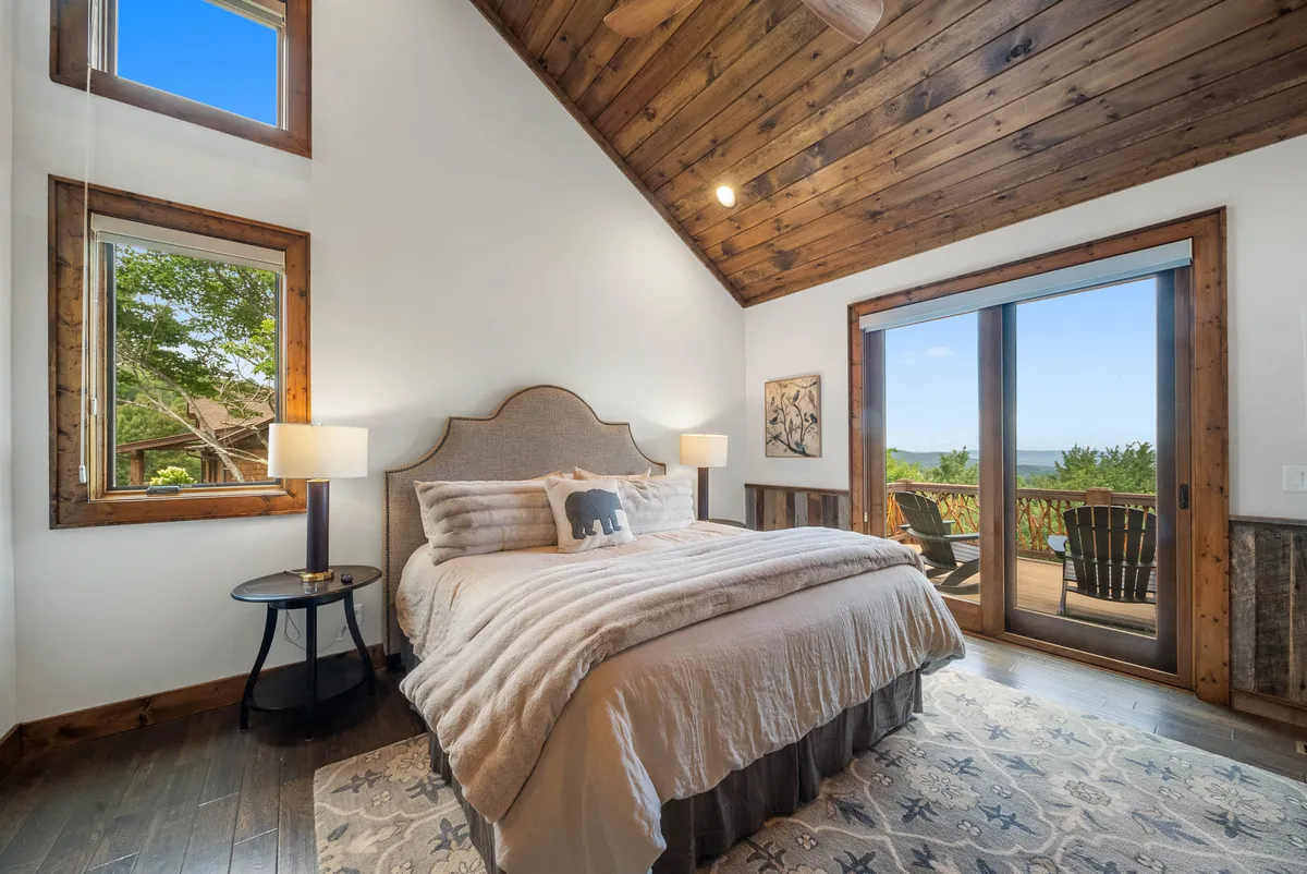 A serene king bedroom featuring a plush bed with premium bedding, large windows framing stunning mountain views, a wooden ceiling, and an art piece. Soft, natural light fills the room, creating a tranquil retreat perfect for relaxation.