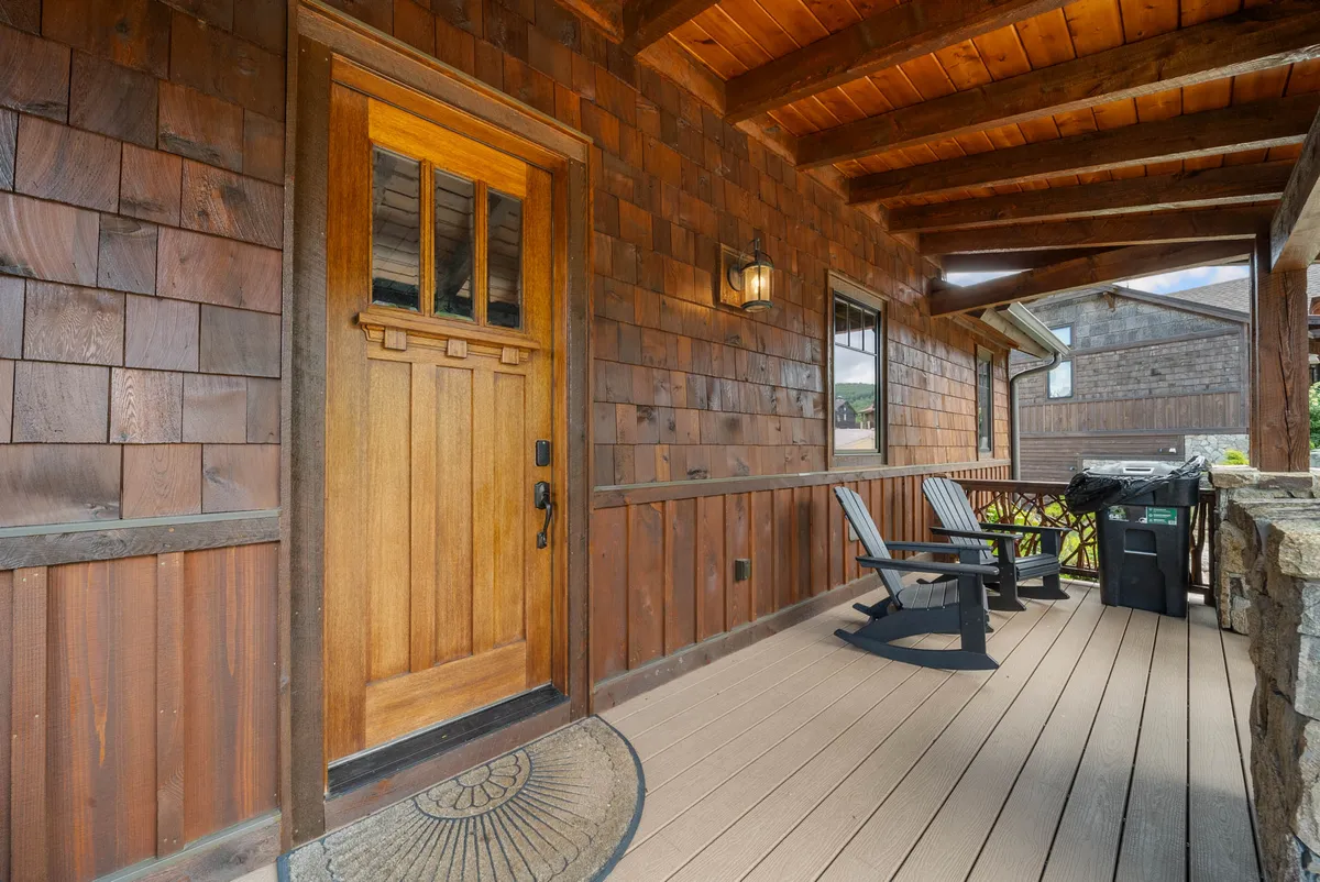 The image shows the inviting exterior of the property, featuring a wooden door framed by a cozy porch. Two classic rocking chairs are arranged beside an outdoor grill, suggesting relaxation and al fresco dining. The porch overlooks the natural mountain landscape, capturing the tranquil atmosphere and stunning sunrise views, perfect for couples seeking a romantic retreat.