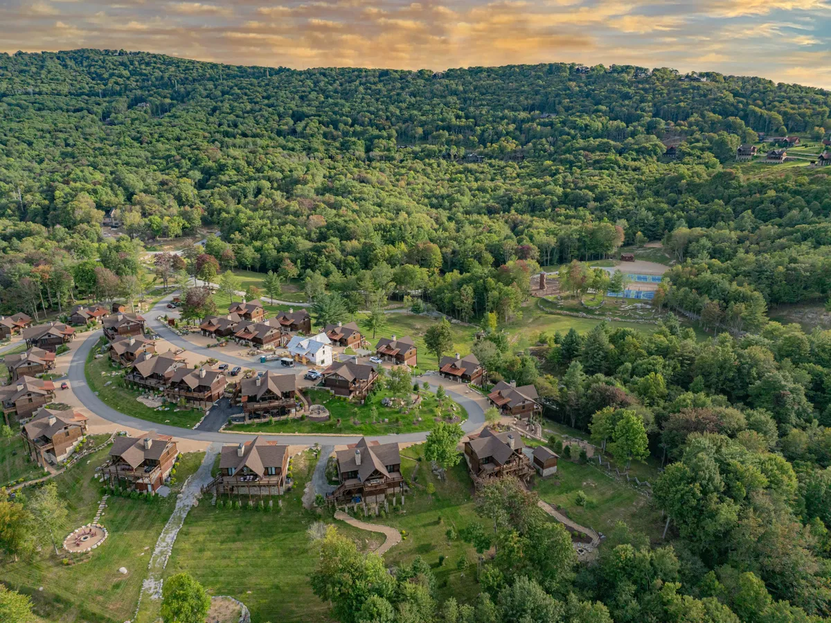 This image features a circular arrangement of cozy cabins surrounded by lush greenery, with visible playground and basketball court areas nearby. Families are seen gathering, children are playing outdoors, and the scene is set during a beautiful sunset, hinting at the vibrant community life and resort-style amenities available within the enclave.