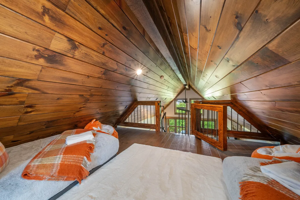 A cozy loft bedroom with orange-accented bedding, open wooden beams, and an open railing that leads the eye toward the outdoors. The visual cue of the outdoors just beyond the railing highlights the indoor-outdoor lifestyle.