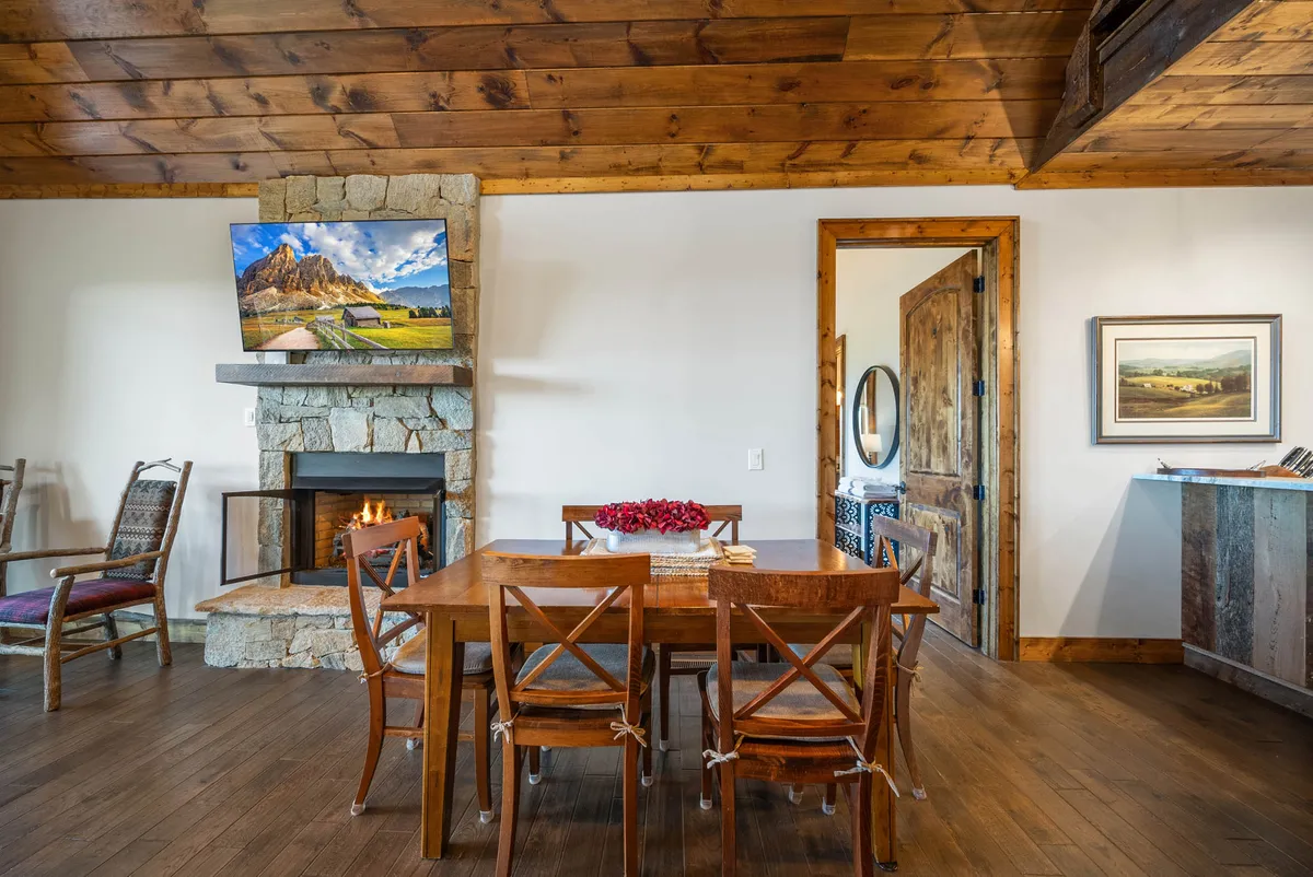 A broad view of the living area featuring a wooden dining table set for group gatherings, a prominent stone fireplace with a warm fire, a television, and a high wooden ceiling. The inviting arrangement suggests both intimate family dinners and cozy group activities during chilly evenings.